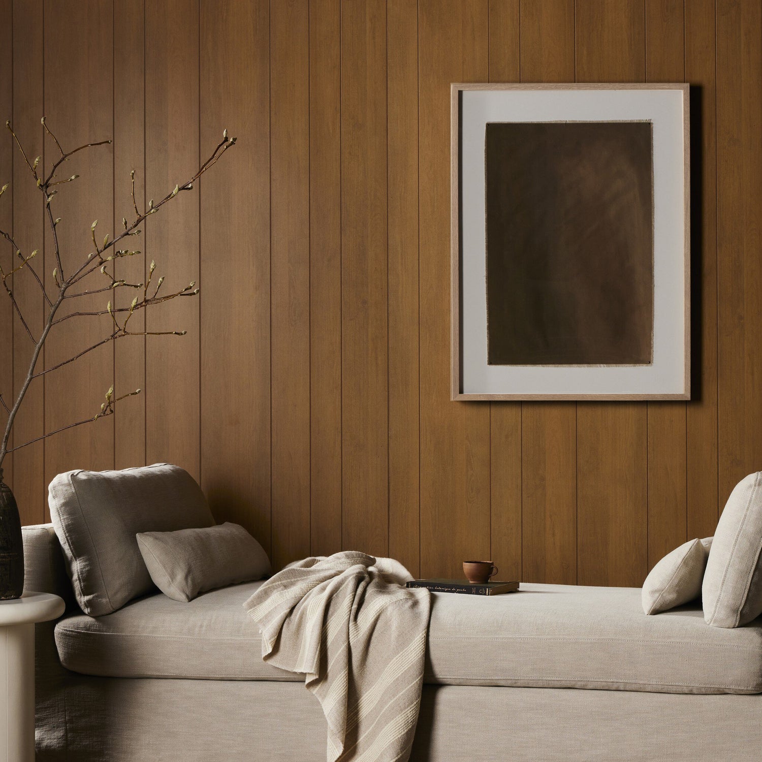 Serene living room with wood paneling, featuring a framed abstract artwork in earthy tones, a comfortable beige chaise lounge, and a decorative branch with buds, showcasing a minimalist and nature-inspired aesthetic