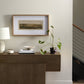 Modern entryway with framed landscape artwork, floating wooden shelf displaying books, plant, and decorative items, and a partial view of a staircase, showcasing a blend of natural elements and minimalist design